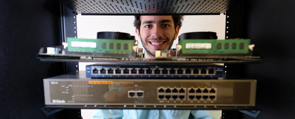 Student smiling into camera through a rack of electronics.