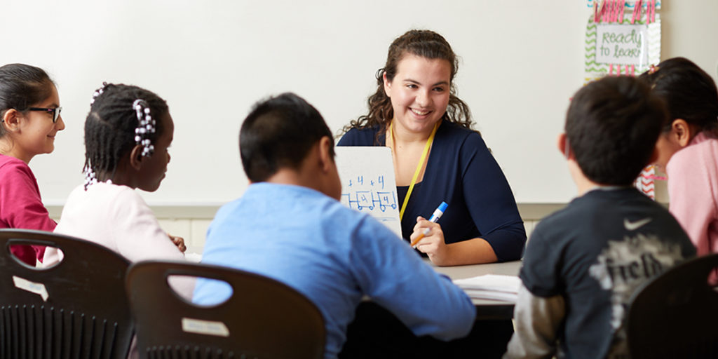 The photo depicts a UMW graduate as a professional, instructing students in a classroom setting.