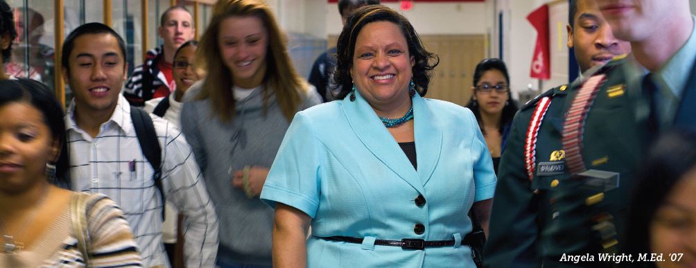 This image depicts a UMW M.Ed. graduate standing in the high school hallway where she teaches in between classes, surrounded by students.