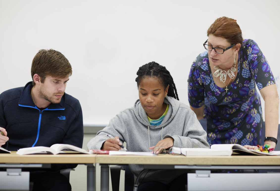 Two students and a professor looking at books on a desk.