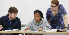 Two students and a professor looking at books on a desk.