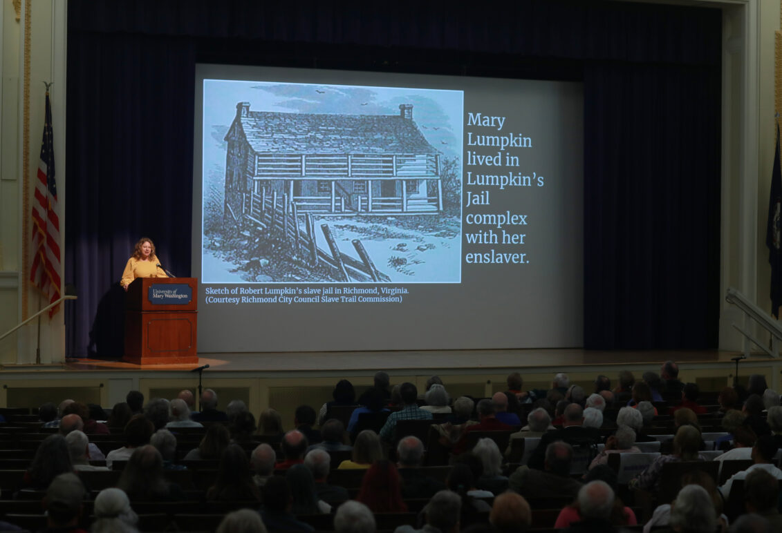 Presenter standing behind podium with a slide behind her. The slide is a sketch of a jail with the text, "Mary Lumpkin lived in Lumpkin's Jail complex with her enslaver".