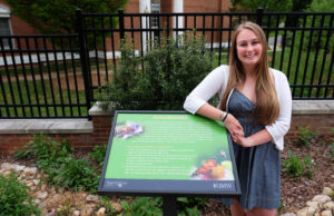 Senior biology and environmental sciences major Maggie Magliato poses with a sign from the pollinator walk she created on campus. Photo by Norm Shafer.