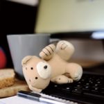 computer keyboard with coffee, food and teddy bear nearby