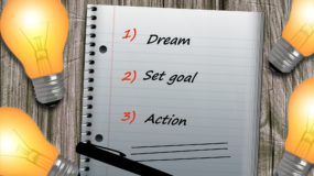 notepad surrounded by lightbulbs. notepad reads a list; dream, set goal, action.