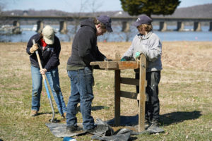 Olivia Larson (left) digs in the dirt, while Matthew Pang Delaney Resweber (far right) use a shaker screen to search for artifacts that could give clues to the location of a lost Patawomeck village. Photo by Suzanne Carr Rossi.