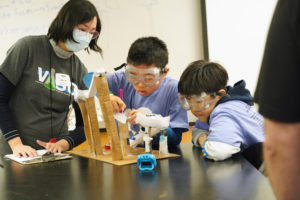 James Pijai (center) and Seungwoo Oh (right), both 10, make adjustments after receiving feedback from Event Supervisor Julia Yao. Competitors in the 'Roller Coaster' category were allowed only two gaps and a loop to slow the journey of a ball along the track they built. The target time for the ball to make it to the end of the track was 40 seconds. Photo by Suzanne Carr Rossi.