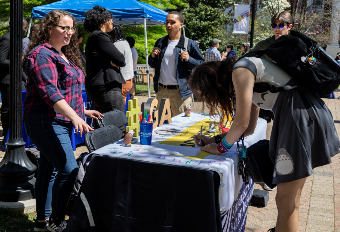A student bends over a table to write something while other students surround the table, which is topped with an "#SGA" decoration