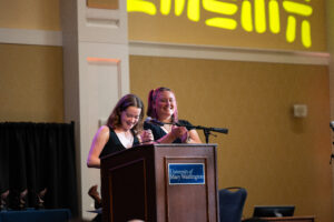 The women in black, seniors Faith Jones and Fiona Archer, captivated the audience with a series of corny jokes produced through 'SammyChatBot,' spreading laughter that set the tone for the evening. Photo by Carson Berrier