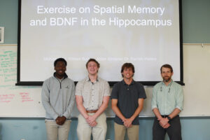 Junior biological science majors William Burkey, Aidan Kimener and Michael Afful pose with Professor Parrish Waters in front of their presentation on whether exercise improves memory. Photo by Karen Pearlman.