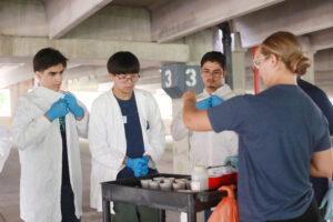 Hannah Steele '26, who earned a Noyce Scholarship at UMW, demonstrates to high schoolers Zubair Mohammadzai (left), Michael Young (center) and Maiwand Barakzai (right) how to soak the cotton balls in washable paint before throwing them at black canvases hanging in UMW's Alvey Parking Deck. Photo by K Pearlman Photography.