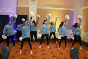 Members of UMW Pep Dance, wearing light blue UMW sweatshirts, black tights and sneakers, perform a cheer with white pom-poms.