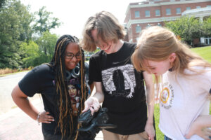 Three college students, one who recently graduated, gather around a video camera to view footage they just shot.