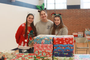 Three University of Mary Washington students stand indoors at the Walker-Grant Center in front of stacked, wrapped gift boxes prepared for preschool children.