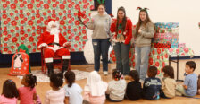Three University of Mary Washington students stand next to Santa in front of a preschool classroom while assisting with the distribution of gift boxes.