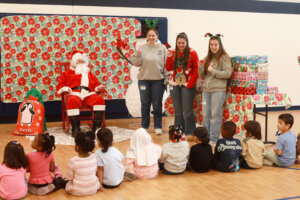 Three University of Mary Washington students stand next to Santa in front of a preschool classroom while assisting with the distribution of gift boxes.