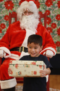 A smiling preschool boy holds a wrapped gift box while Santa stands in the background inside a decorated room.