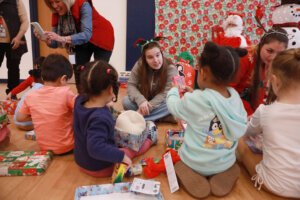 A University of Mary Washington student sits on the floor with preschool children as they open gift boxes in a decorated room at the Walker-Grant Center.