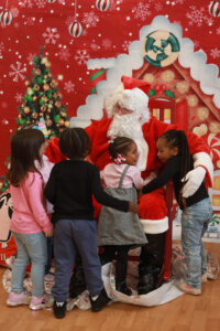 A group of preschool children hug Santa, portrayed by an adult in a red suit, inside a classroom decorated for a holiday celebration.