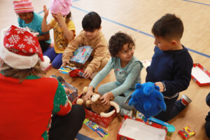 Preschool children sit on the floor opening wrapped gift boxes inside a classroom during a holiday delivery event.