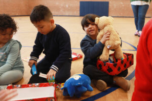 A preschool child hugs a teddy bear while other children nearby hold toys and smile inside a classroom.