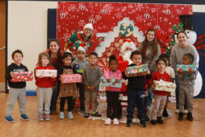 A group of University of Mary Washington students and a staff member pose with Santa and a classroom of preschool children holding gift boxes inside the Walker-Grant Center.