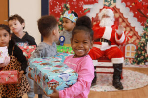 A smiling preschool girl wearing pink holds a wrapped gift box inside a classroom.