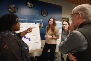 A group of students stands in the Jepson Science Center lobby while discussing a research poster about invasive species and native mussels.