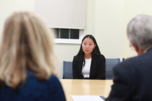 A college student in a suit answers a question of two judges with their backs to the camera.