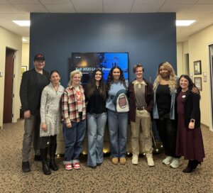A group of student interns, mentors and staff stand together in the University Communications office during a video production showcase.