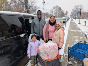Adult woman and family members stand with bag of wrapped gift boxes during a pickup at a campus location.