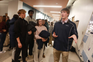 William Burkey presents research in front of a poster while students Tucker Burkey, Demetrious Walker, Audrey Sivak and Hannah Siraj listen.