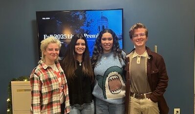 Four student interns, one junior and three seniors, stand together in the University Communications office during a video production showcase.