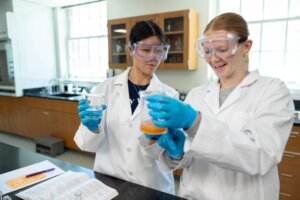 Two college students wearing lab coats, gloves and goggles look at a beaker filled with liquid in a science lab at the University of Mary Washington