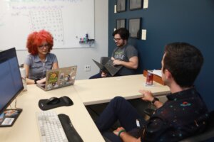 Students and a professor sit in the professor's office while working on laptops and collaborating on research
