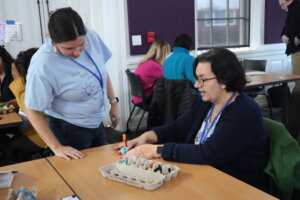 A lady sits at a table choosing from an array of colorful objects nestled into an egg carton, while Maker Mindset Conference originator Kristina Peck looks on.