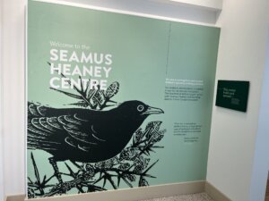 A mural at the Seamus Heaney Centre at Queen's University in Belfast. Seafoam green background with a blackbird sitting on branches. Text reads: Welcome to the Seamus Heaney Centre. We aim to strengthen and expand Belfast's dynamic writing practices. Our emblem, the blackbird, is inspired in part by the ancient Irish poem 'The Blackbird of Belfast Lough,' which both Seamus Heaney and founding director Ciaran Carson translated. Italicized quote: 'There are, to paraphrase Wallace Stevens, at least thirteen ways of looking at a blackbird; and the blackbird can be heard in many ways... Poetry resides in that ambiguity.' - Ciaran Carson. 