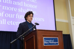 A student singing the Mary Washington Alma Mater at a podium with the lyrics on a screen behind him.