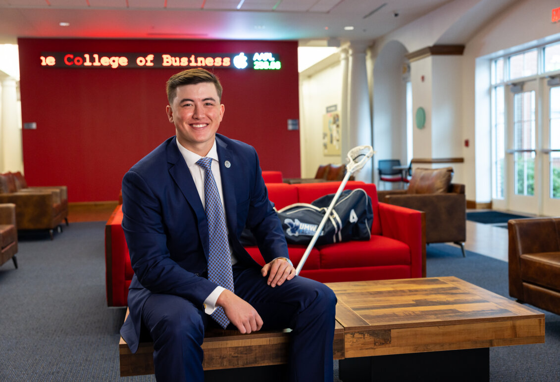 Photo of Jay Hooker in a business suit holding a lacrosse stick in the lobby of the College of Business