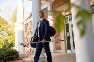 Photo of UMW senior Jay Hooker dressed in a business suit with a lacrosse bag on his shoulder and holding a lacrosse stick while exiting the College of Business on campus