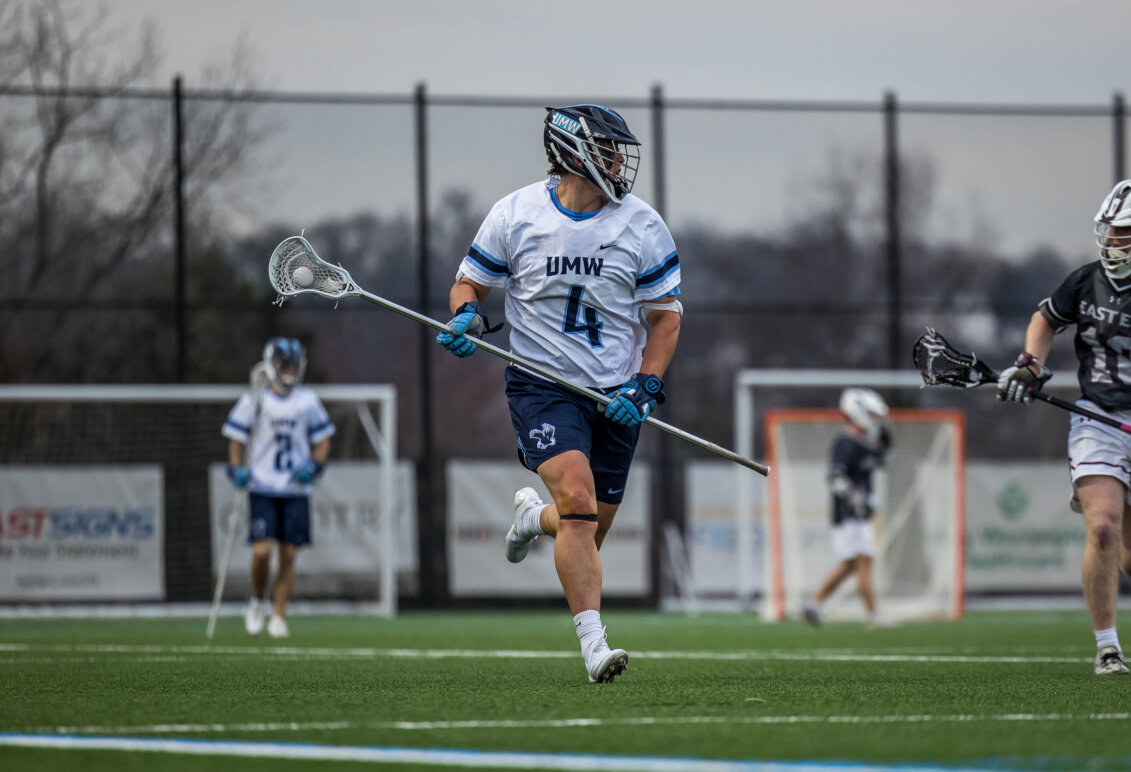 Photo of a Jay Hooker in a white jersey playing lacrosse for UMW holding a lacrosse stick.