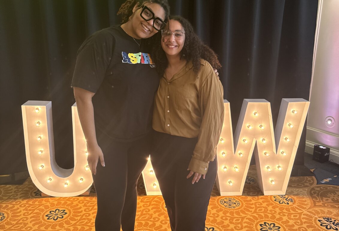 Two women hug and smile for the camera onstage in front of lit-up "UMW" letters