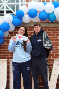 Two students with an 'I love Mary Wash' bumper sticker and a 'Go Wash' foam finger under a balloon arch.