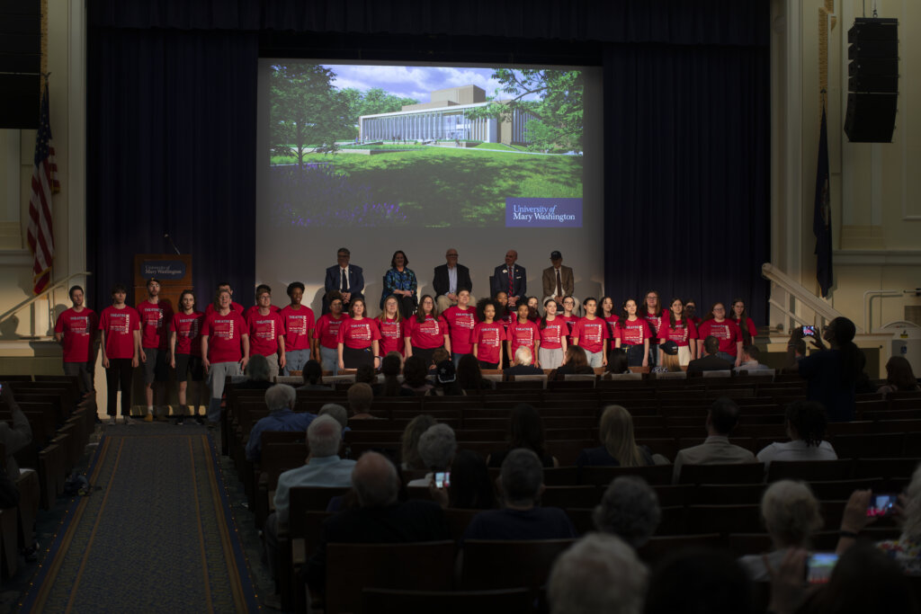 A group of students dressed in red stands in front of a rendering of a new theatre building