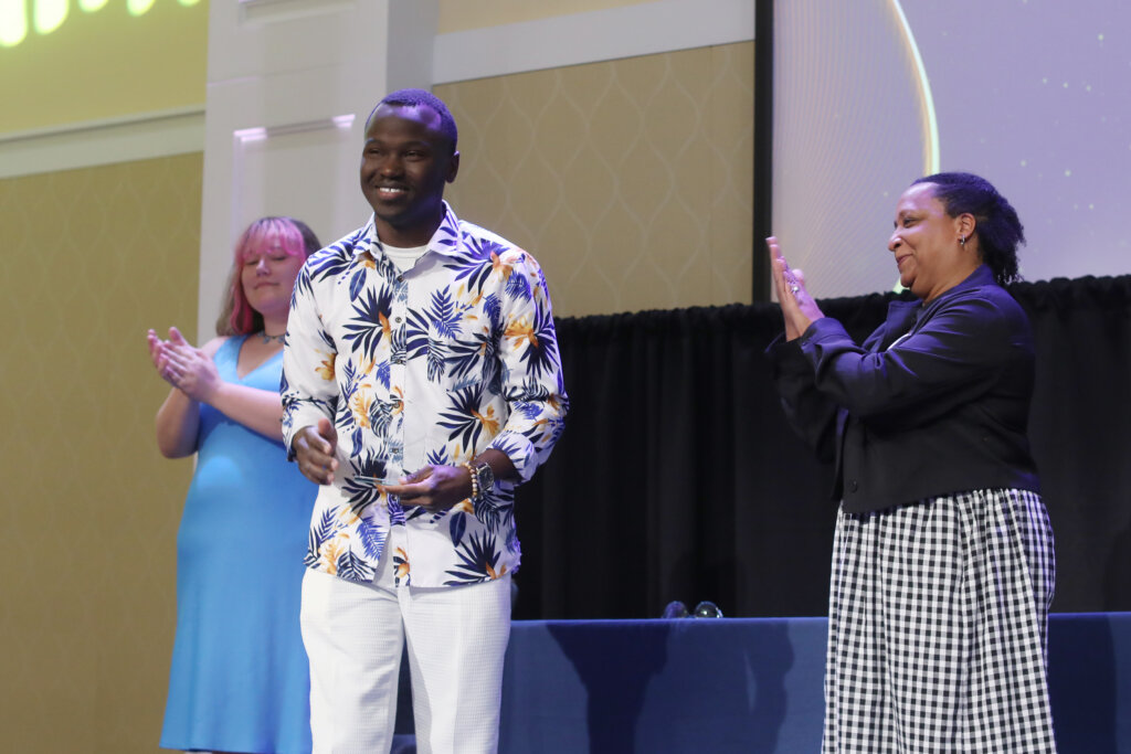 Three people stand onstage, one of whom is holding an award and two who are clapping.