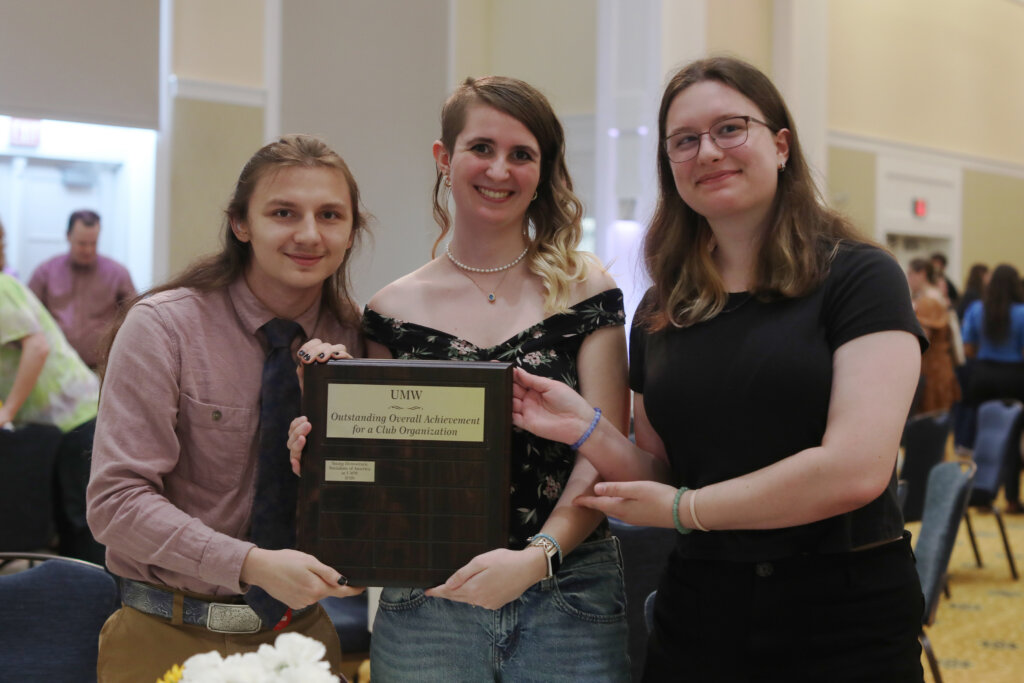 Three students stand in a crowded room holding an award.