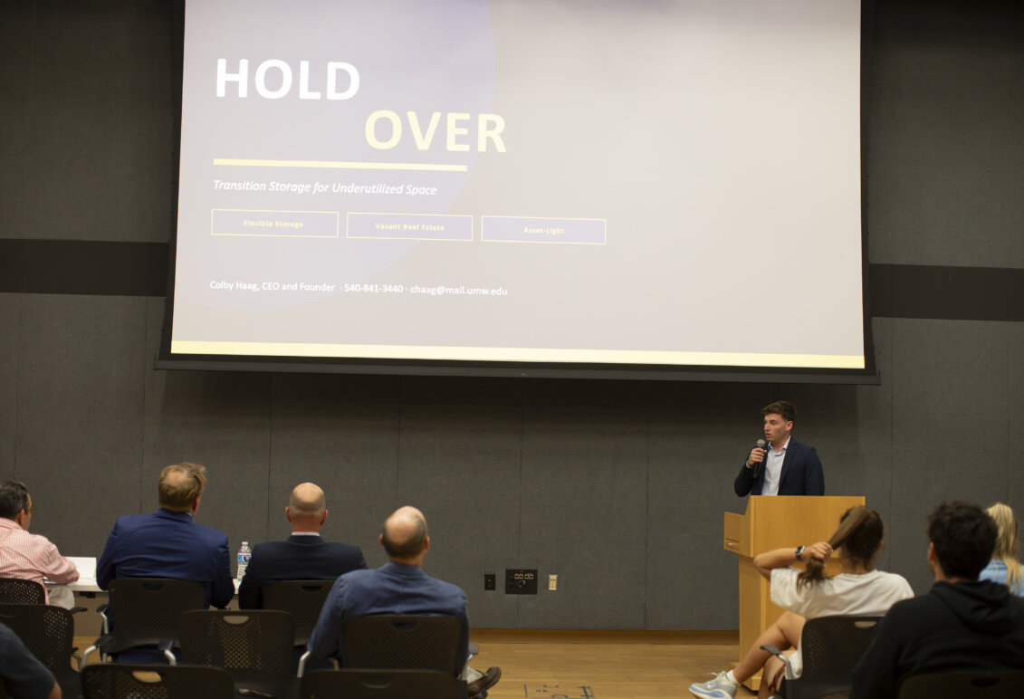 A guy holding a microphone behind a podium in front of a screen that says "Hold Over."