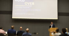 A guy holding a microphone behind a podium in front of a screen that says "Hold Over."