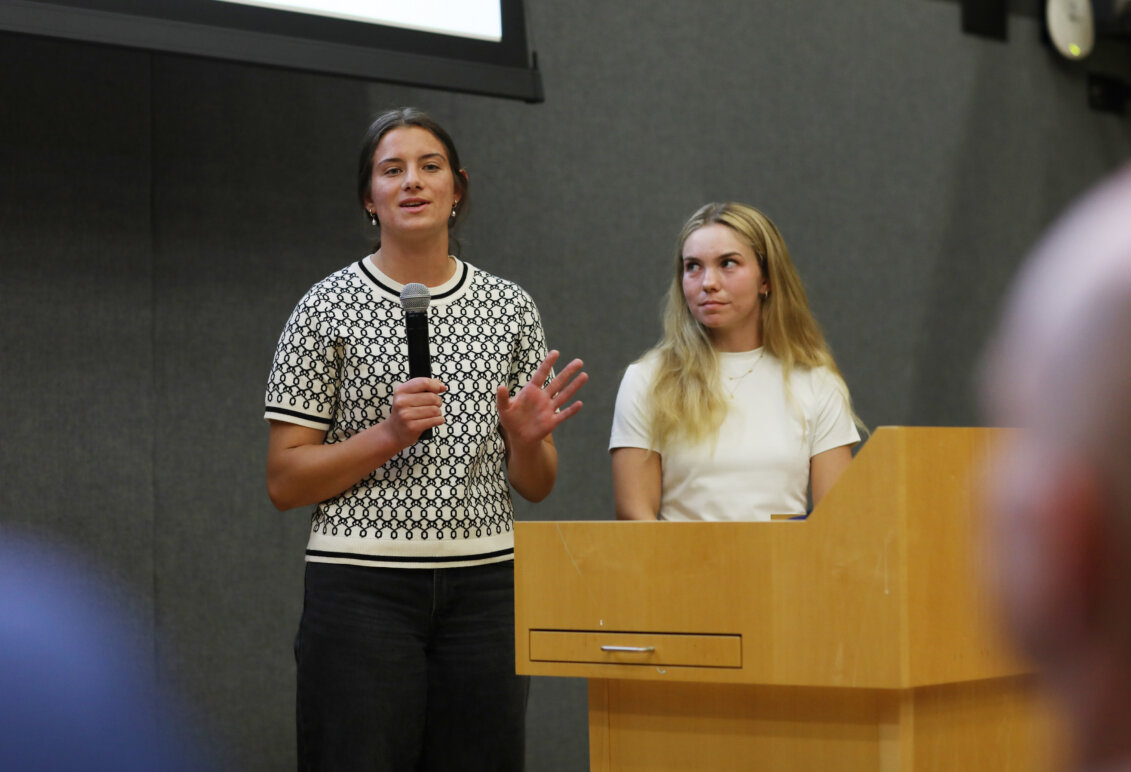 Two girls stand behind a podium, while one speaks into a microphone