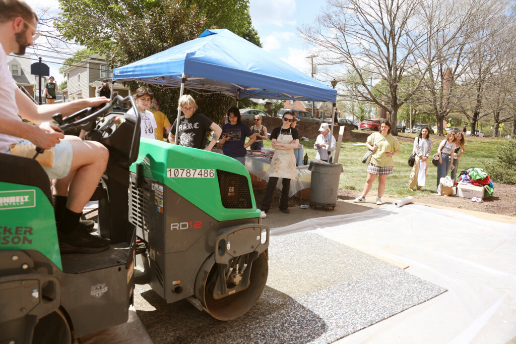 A professor rides the steamroller while students watch from a small tent and a line next to the tent.